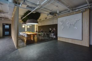 Room in a loft style. There are light and dark tables, light and dark chairs. There is stand for office supplies between light tables. Above them hang large lamps with artificial leaves. A few laptops are on the table. On the right there is a wall with world map on it. On the left there is an ornamental wooden partition. At the top you can see the communications, lamps, speakers and a projector. Also at the end of the room there are doors. Place for coworking.